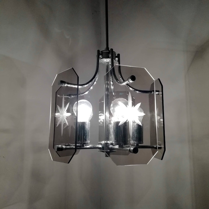 May include: A vintage chandelier with a chrome frame and three light bulbs. The fixture features gray, geometric glass panels with etched starburst designs. The light fixture is suspended from a black rod.
