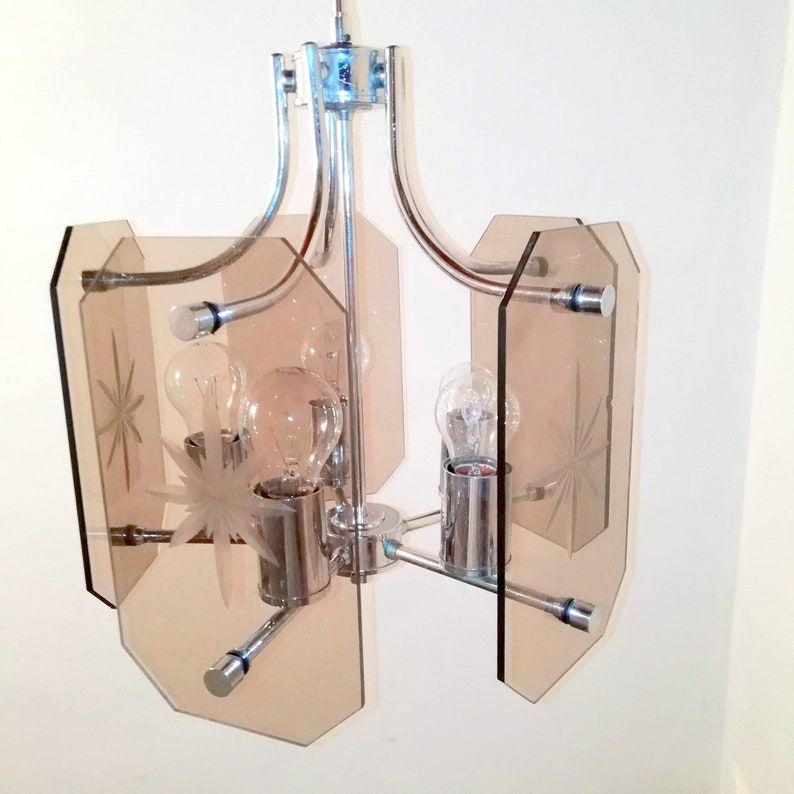 May include: A vintage chrome and glass chandelier with four light bulbs. The chandelier features a starburst design etched into the amber-colored glass panels. The fixture has a mid-century modern aesthetic.
