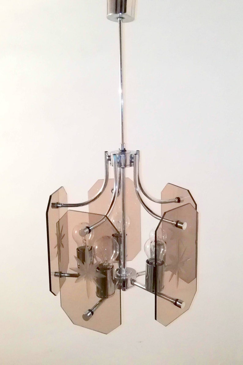 May include: A chrome and glass chandelier with a mid-century modern design. The fixture has a central stem and curved arms holding light bulbs. The light shades are a light brown color and have a starburst design etched into the glass.