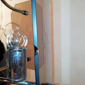 May include: A close-up of a decorative mirror with a black frame. The mirror has a floral design etched into the glass. The frame has a broken section on the edge. A light fixture is visible in the background.