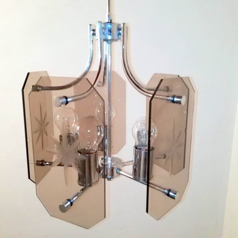 May include: A vintage chrome and glass chandelier with four light bulbs. The chandelier features four light brown glass panels with starburst designs. The fixture has a polished chrome finish and is suspended from a single point.