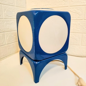 May include: A retro blue cube lamp featuring two large, white circular insets. The lamp sits on a matching blue base with curved legs. A cream-coloured cord extends from the base. A stylish, geometric lighting fixture.