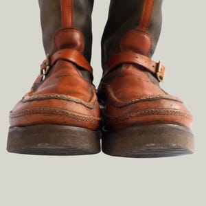 May include: Pair of brown leather boots with a dark green upper. The boots feature a buckle strap across the top of the foot and a thick, dark brown sole. The leather has a worn, vintage appearance.