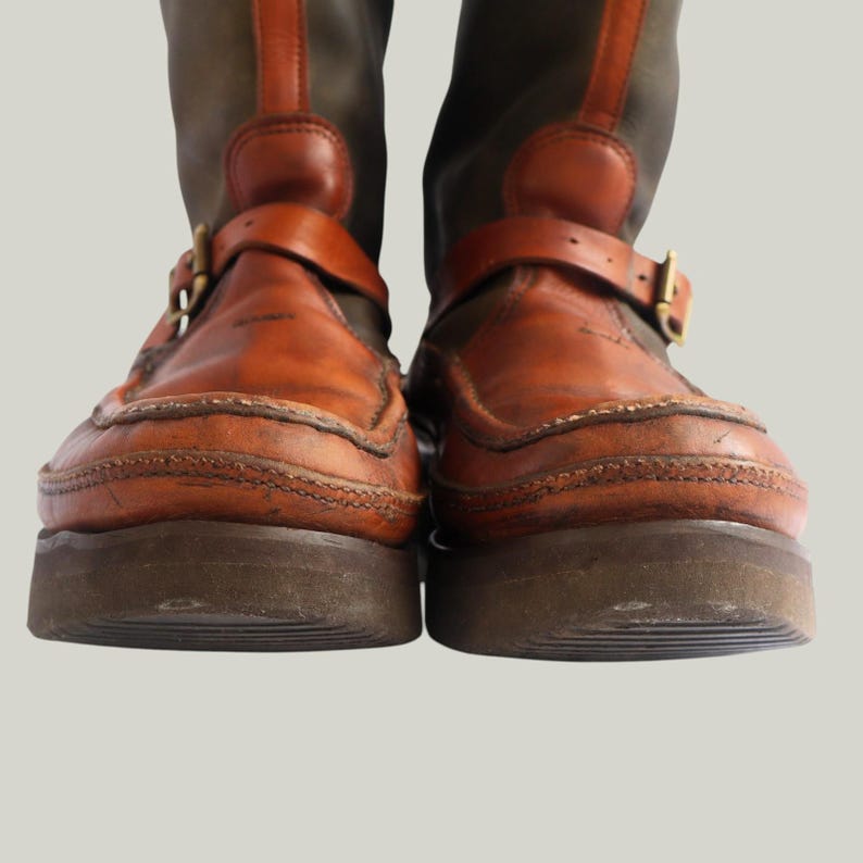 May include: Pair of brown leather boots with a dark green upper. The boots feature a buckle strap across the top of the foot and a thick, dark brown sole. The leather has a worn, vintage appearance.