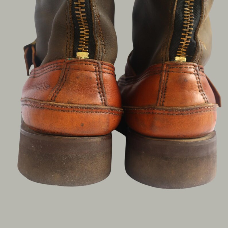 May include: Close-up of two brown and tan leather boots. The boots have a zipper up the back with a gold-colored zipper. The soles are thick and dark brown. The leather has a worn appearance.