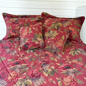 May include: A quilted bedding set featuring a deep red base with a dense floral pattern in shades of pink, yellow, and green. The set includes a large rectangular quilt, two rectangular shams, and two square throw pillows, all with matching fabric.