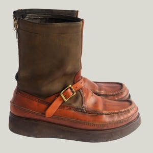 May include: A pair of brown and orange leather boots with a platform sole. The boots have a zipper on the side and a buckle strap. The upper part of the boot is a darker brown, while the lower part is a lighter orange.