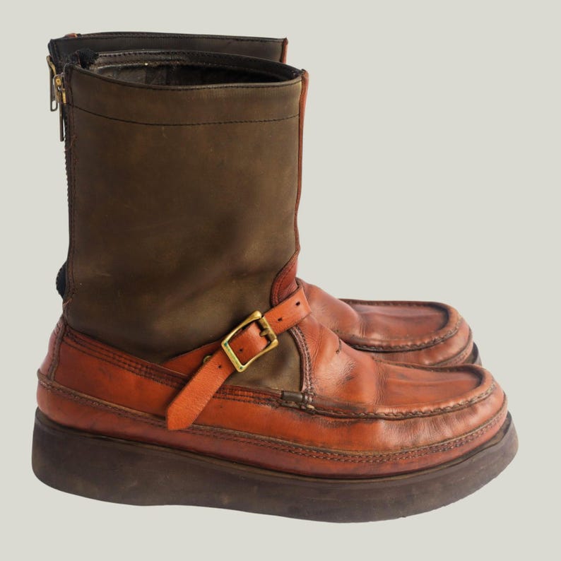 May include: A pair of brown and orange leather boots with a platform sole. The boots have a zipper on the side and a buckle strap. The upper part of the boot is a darker brown, while the lower part is a lighter orange.