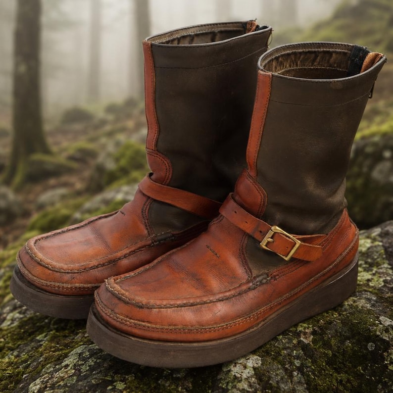 May include: A pair of brown and tan leather boots with a rugged design. The boots feature a rounded toe, a strap with a gold-colored buckle, and a thick sole. The boots are approximately 10 inches tall.