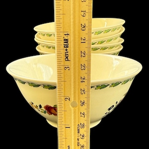 May include: A stack of cream-coloured ceramic bowls featuring a green leaf vine pattern along the rim and floral motifs. A wooden ruler is placed vertically in front, showing the front bowl has a diameter of approximately 19 cm.