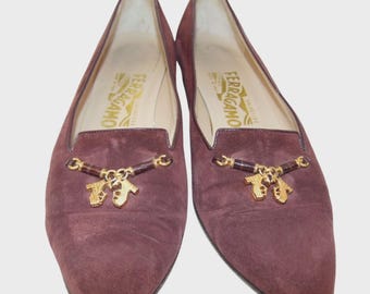 May include: A pair of burgundy suede loafers with a pointed toe. The shoes feature a gold-tone embellishment with a horse-bit design and the brand name "Ferragamo" printed on the insole.