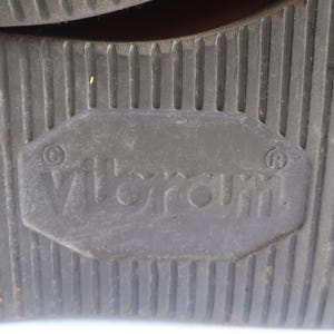 May include: Close-up of a dark gray shoe sole with a textured, ridged pattern. The sole features the brand name "Vibram" in a rectangular inset. The sole is designed for traction and durability.