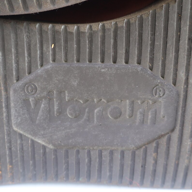 May include: Close-up of a dark gray shoe sole with a textured, ridged pattern. The sole features the brand name "Vibram" in a rectangular inset. The sole is designed for traction and durability.