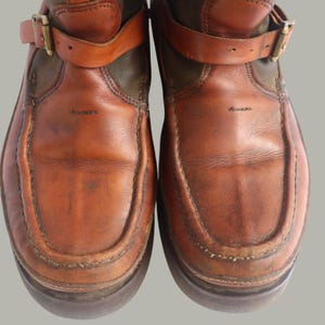 May include: Close-up of a pair of brown leather boots. The boots have a distressed appearance with visible wear and tear. They feature a strap with a brass buckle and a dark green fabric detail at the top. The soles are dark brown.