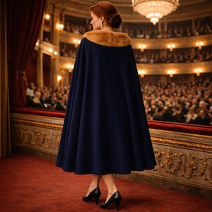 May include: A navy blue cape with a tan fur collar. The cape is worn over a black dress and black heels. The person is standing on a red carpet in an opera house, facing away from the camera.