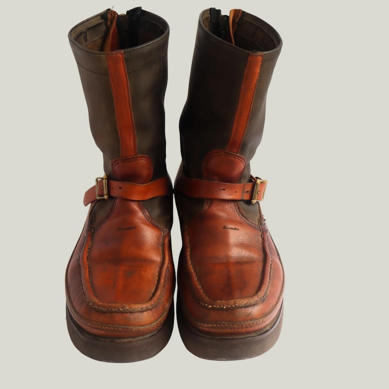May include: A pair of brown and orange leather boots with a zipper closure and buckle straps. The boots have a round toe and a thick sole. The upper portion of the boots is a darker brown, with orange accents and a leather strap around the ankle.