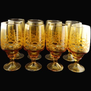 Puede incluir: Una colección de ocho copas de vino de color ámbar. Cada copa presenta un patrón decorativo repetitivo en un tono ámbar más claro. Las copas tienen forma de cuenco redondeado y se asientan sobre un tallo corto y una base circular. El fondo es negro.