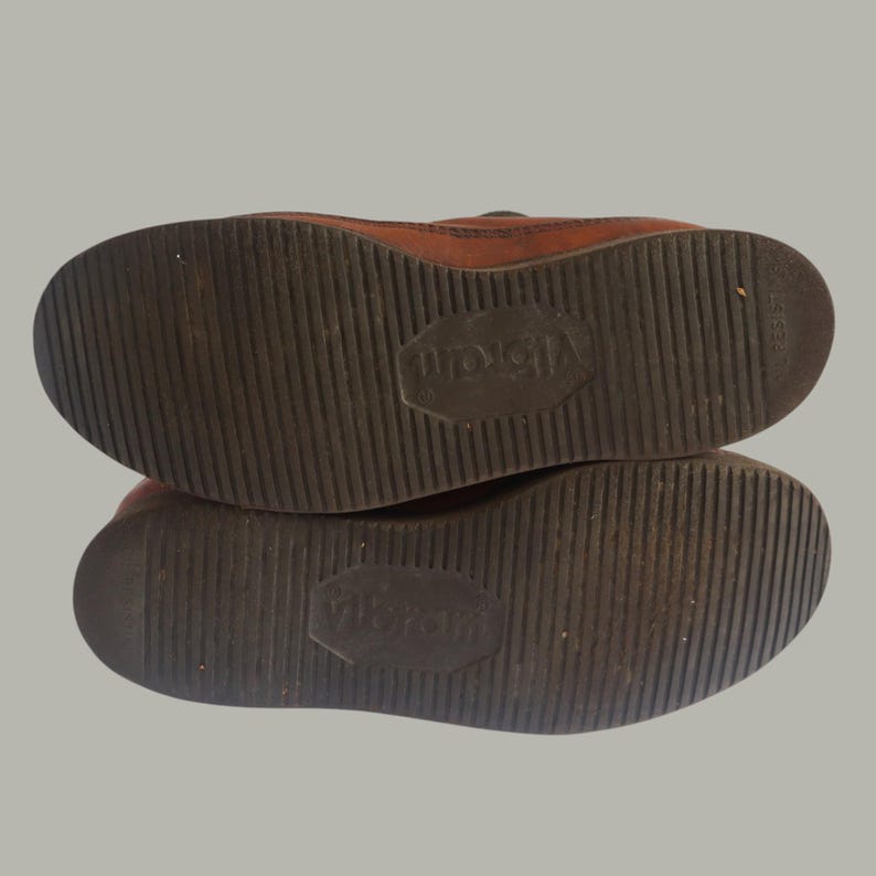 May include: Close-up of the soles of two brown leather shoes. The soles are dark brown with a textured, ridged pattern. The brand name "Vibram" is embossed on each sole. The shoes appear to be well-worn.