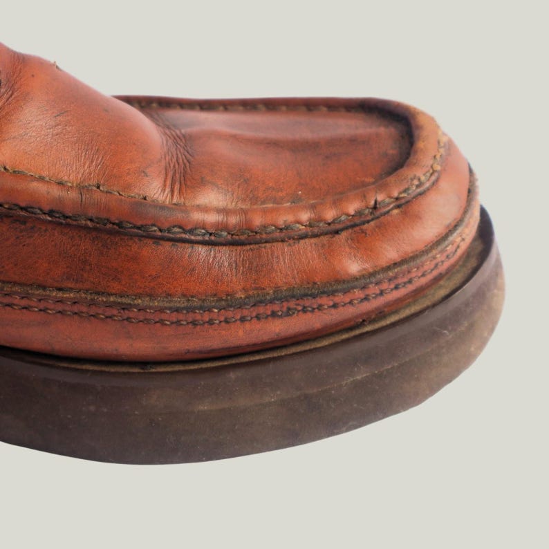 May include: Close-up of a brown leather shoe. The shoe features a moccasin-style design with visible stitching and a thick, dark brown sole. The leather has a worn, textured appearance.