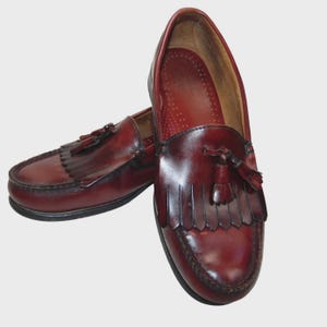 May include: A pair of burgundy leather loafers with decorative tassels. The shoes have a classic design with a rounded toe and a low heel. The leather has a polished finish, and the soles are black.
