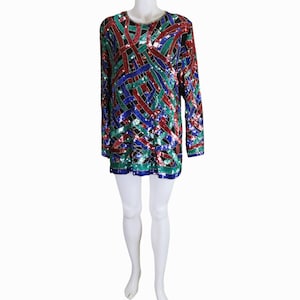 Puede incluir: Una blusa de manga larga estilo túnica con un diseño de lentejuelas de colores. El patrón presenta líneas que se cruzan en rojo, verde, azul y negro. El escote y el dobladillo están adornados con un borde contrastante. La blusa se muestra en un maniquí blanco.