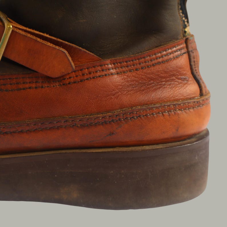 May include: Close-up of a brown leather boot with orange and dark brown sections. The boot features a buckle and strap detail, with visible stitching and a thick, dark brown sole. The leather shows signs of wear.