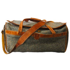 May include: A vintage duffel bag with a gray tweed body and brown leather accents. The bag features a top handle, a shoulder strap, and a small lock. The leather is a warm brown color, and the bag has a classic, retro style.