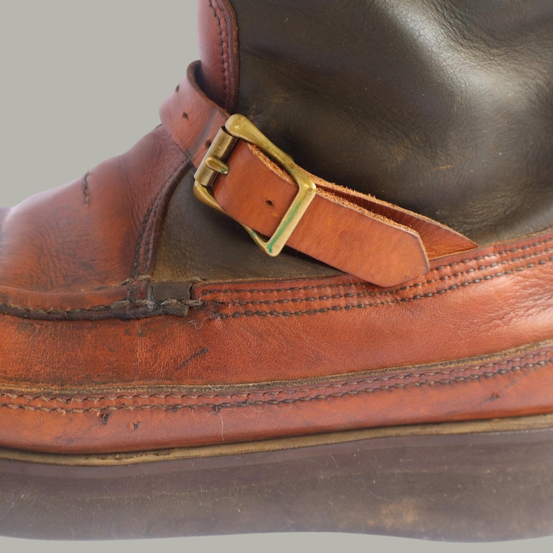 May include: Close-up of a brown leather boot with a buckle. The boot features a reddish-brown leather lower section and a darker brown upper section. A leather strap with a gold-colored buckle is fastened across the boot's ankle area.