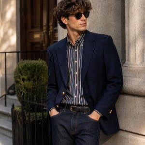 May include: A navy blue blazer with gold buttons, a dark button-down shirt featuring vertical white and red stripes, and dark blue denim jeans with a black leather belt. The outfit is completed with dark-framed sunglasses and wavy brown hair.