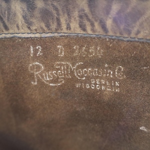 May include: Close-up of the interior of a brown leather boot. The text "12 D 2656" is visible, along with the brand name "Russell Moccasin Co." and the location "Berlin Wisconsin" in white lettering.