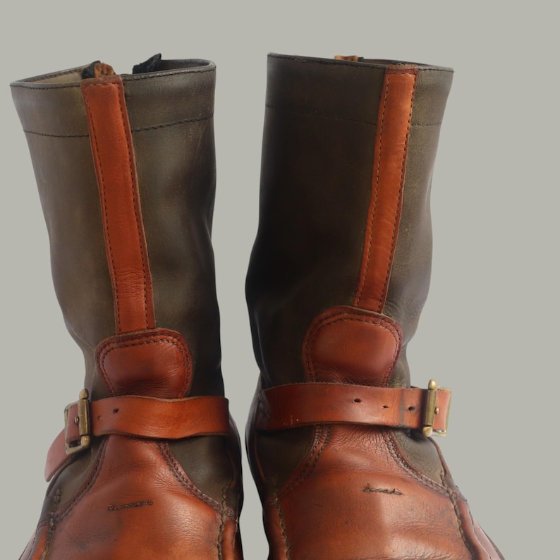May include: Pair of brown and olive green leather boots. The boots have a mid-calf height with a buckle strap around the ankle and a vertical orange leather stripe down the back. The boots have a zipper closure and a round toe.