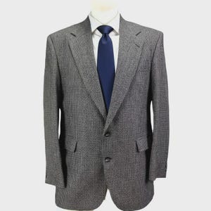 Può includere: Un blazer grigio con un sottile motivo strutturato, con una classica chiusura a due bottoni e revers a lancia. Il blazer è abbinato a una camicia bianca e una cravatta blu navy. La giacca ha due tasche anteriori.