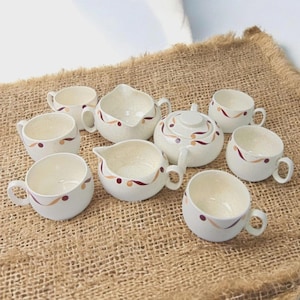 May include: A cream-colored tea set with a speckled finish, featuring a teapot, cups, and creamers. Each piece is adorned with a decorative pattern of burgundy and gold swirls. The set is arranged on a burlap surface.