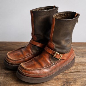May include: A pair of brown leather boots with a rugged, worn appearance. The boots feature a mid-calf height, a buckle strap, and a thick sole. The upper portion is a darker brown, contrasting with the lighter brown lower section.