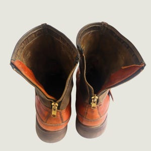 May include: Pair of brown and orange leather boots with open zippers. The boots have a rugged, worn appearance, with visible stitching and a brass zipper. The interior lining is a contrasting orange color, and the boots appear to be well-constructed.