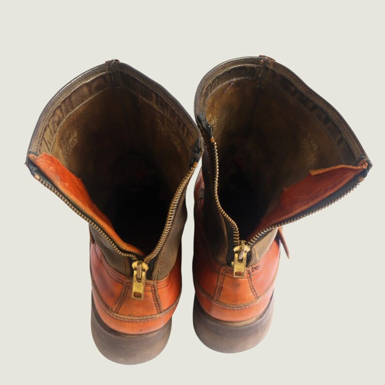 May include: Pair of brown and orange leather boots with open zippers. The boots have a rugged, worn appearance, with visible stitching and a brass zipper. The interior lining is a contrasting orange color, and the boots appear to be well-constructed.