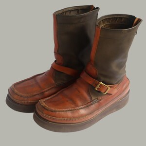 May include: A pair of brown leather boots with a platform sole. The boots have a dark brown upper with orange accents and a buckle strap. The soles are a darker brown color.
