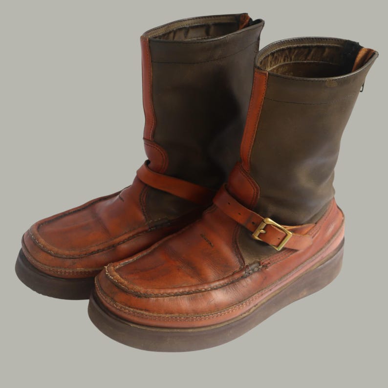 May include: A pair of brown leather boots with a platform sole. The boots have a dark brown upper with orange accents and a buckle strap. The soles are a darker brown color.