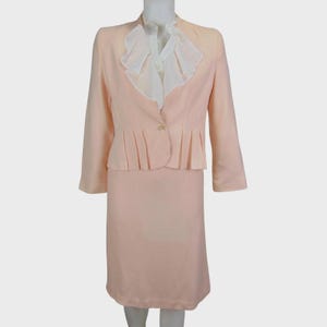 Könnte beinhalten: Hellpfirsichfarbener Hosenanzug mit passendem Blazer und weißer Bluse. Der Blazer hat einen einzelnen Knopf und einen plissierten Bund. Die Bluse hat einen Rüschenkragen. Klassischer Stil.