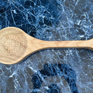 Pode incluir: Uma colher de madeira clara com um design de mandala decorativo esculpido na tigela. A colher tem um cabo longo e um buraco na extremidade para pendurar. A colher tem aproximadamente 30 cm de comprimento.