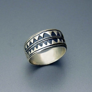 Puede incluir: Un anillo de plata con un diseño geométrico. El anillo presenta una banda con una serie de formas triangulares en un patrón repetitivo. El anillo tiene un acabado pulido y está sobre un fondo gris.