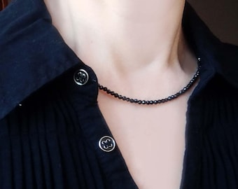 May include: A delicate necklace composed of small, faceted black beads worn around the neck. The necklace is paired with a black collared shirt featuring three visible black buttons with silver-toned rims along the front placket.
