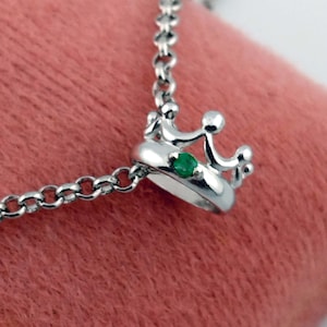 May include: A silver crown pendant necklace with a small green gemstone. The crown is detailed with rounded points and a circular base. The necklace chain is a delicate link style, and the background is a soft pink.