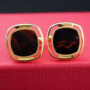Peut inclure: Une paire de boutons de manchette dorés avec un cadre carré aux coins arrondis. Chaque bouton de manchette contient une incrustation centrale en pierre brun foncé et noire avec des motifs naturels tourbillonnants, sertie dans un bord métallique poli.