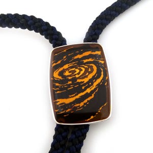 Op de afbeelding: Een bolo-stropdas met een rechthoekige hanger met een wervelend oranje en zwart ontwerp, gezet in een zilveren frame. Het gevlochten koord is marineblauw en zwart. Dit accessoire voegt een unieke touch toe aan elke outfit.