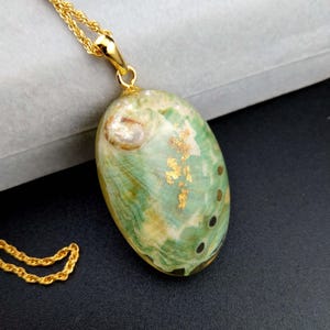 May include: An oval pendant necklace featuring a polished abalone shell with swirling green, white, and tan patterns, accented by embedded gold leaf flakes. The pendant is suspended from a gold-toned twisted rope chain with a polished metal bail.