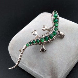 Puede incluir: Un broche de lagarto en tono plateado con una hilera de piedras redondas de color verde vibrante a lo largo de la espalda y la cola. El lagarto tiene un cuerpo metálico texturizado, cuatro patas extendidas y una pequeña piedra roja como ojo.