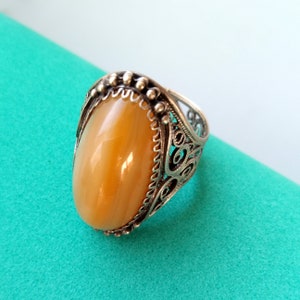 Pode incluir: Um anel de prata com uma pedra grande, lisa e oval de cor âmbar, engastada numa moldura de metal decorativa. O aro e a cravação são trabalhados com padrões intrincados de filigrana aberta e pequenos detalhes de contas na borda.