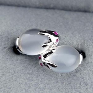 May include: A pair of silver stud earrings. Each earring has a large, oval, milky-white stone, with a silver design and small, round, pink gemstones. The earrings are set against a grey background.