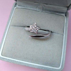 May include: A silver-colored ring with a star-shaped design. The star is adorned with small, clear gemstones. The ring is open, with one end curving around to meet the star. The ring is displayed in a gray velvet box.